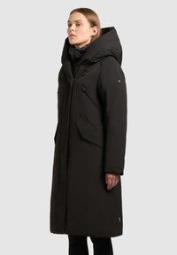 Sort, lang dunjakke med højt krave, flere lynlåslommer og et struktureret stof med en glat finish.