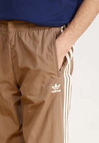 Person klädd i bruna Adidas träningsbyxor med vita ränder och logotyp, hand i det fickan med dragkedja, marina blau skjorta delvis synlig.