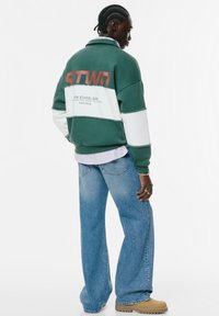 Giacca in fleece color block verde e bianco con logo "STWD", vestibilità rilassata, abbinata a jeans azzurri chiari e stivali beige.