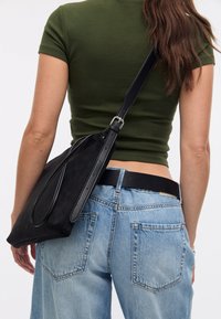 Borsa a spalla nera in tela con una tracolla sottile, design minimalista e texture liscia, indossata con jeans larghi azzurri e una maglietta verde.
