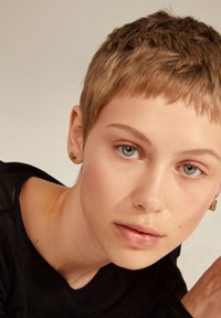Top nero a maniche corte, pelle chiara, capelli biondo texturizzati e corti, occhi azzurro-verdi, indossa orecchini d'oro texturizzati. Sfondo morbido e neutro.