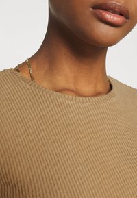 Gros plan sur la partie inférieure du visage et le cou d'une personne portant un pull beige côtelé et un fin collier chaîne en or.