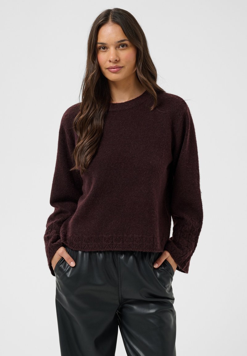 Femme aux longs cheveux bruns portant un pull marron foncé et un pantalon en cuir noir, les mains dans les poches, devant un fond uni.