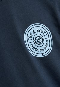 Tissu bleu foncé avec un logo circulaire blanc portant l'inscription « Luke & Stott Born in the Borders » entourant un motif floral.