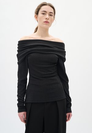 Woman wearing a black off-the-shoulder long-sleeve top with draped neckline and black trousers against a plain white background.