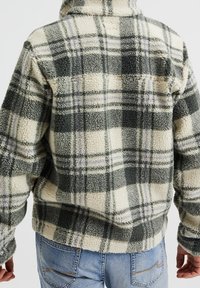 Vihreä-kermanvärinen-laventelivärinen fleece-ruututakki. Pehmeä rakenne, korkea kaulus ja rento istuvuus näkyvin ompeluominaisuuksin. Sinisiä farkkuja käytetty alla.