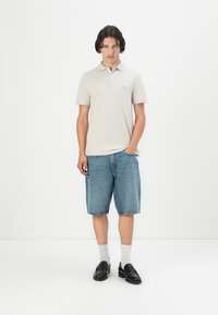 Polo de culoare bej deschis, cu mâneci scurte, asortat cu pantaloni scurți din denim albastru. Loafers negri și șosete albe completează ținuta.