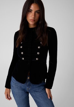 Mujer con cabello largo y oscuro que lleva una chaqueta negra abotonada con botones plateados y vaqueros azules, de pie frente a un fondo gris liso.