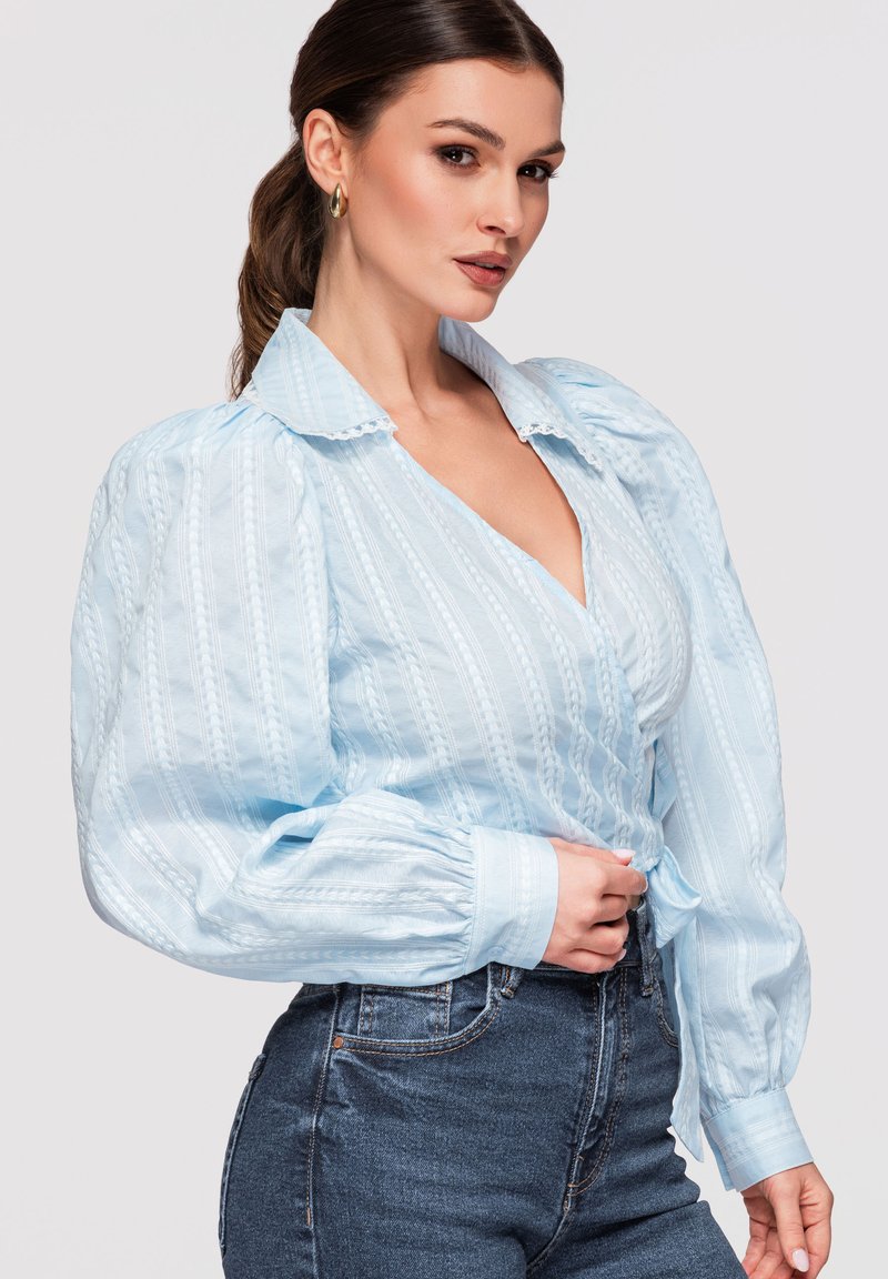Femme aux cheveux bruns tirés en arrière, portant un chemisier bleu clair texturé à manches bouffantes et un jean taille haute foncé, posant avec une main près de la taille.