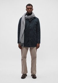 Casaco azul-marinho com botões, um cachecol cinza, calças beiges e botas castanhas. O casaco apresenta um design estruturado e uma textura suave.