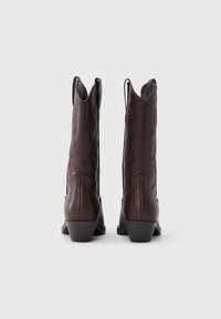 Pár barna bőr cowboy csizma hímzett díszmintákkal, hátulról nézve, áll egy egyszínű világos háttéren.