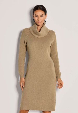Frau in einem beigen, langärmeligen, gerippten Strickkleid mit einem großen Schalkragen und geometrischen Creolen, die vor einem schlichten Hintergrund steht.
