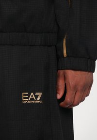 Črn trenirko z teksturirano tkanino, elastičnim pasom in zlatim logotipom EA7 Emporio Armani na spodnjem delu noge. Zlati detajl na rokavu.