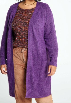 Femme portant un cardigan violet ouvert sur le devant, un haut tricoté multicolore, et un pantalon en velours côtelé marron clair avec une ceinture à nouer.