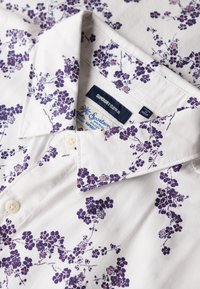 Camisa de manga corta de tejido ligero, con una base blanca y un patrón floral morado. Incluye un cuello con botones y etiqueta de la marca.