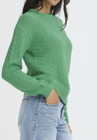 Mujer con un suéter de punto verde texturizado con puños acanalados y jeans de tiro alto azul claro, con una mano metida en el bolsillo delantero.