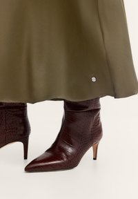 Braune, krokodilpräge Leder-Ankle Boots mit spitzem Zehen und einem 5 cm hohen Stiletto-Absatz, kombiniert mit einem olivgrünen Rock mit silbernem Akzent.