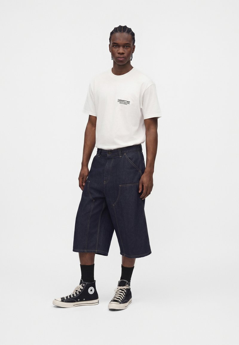Ung mand med fletninger har en hvid kortærmet skjorte, mørke brede shorts til knæet, sorte højtop-sneakers og sorte sokker på.