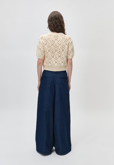 Femme debout de dos, portant un pull en tricot crème à manches courtes et un jean bleu foncé taille haute à jambes larges.