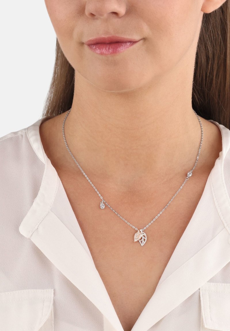 Collana d'argento con un pendente a forma di foglia, caratterizzata da un design texturizzato e una piccola goccia decorativa, indossata sopra una camicia bianca con colletto.