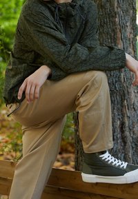 Personne portant une veste verte à motifs, un pantalon Converse beige et des baskets montantes vertes à semelles blanches, assise sur une rambarde en bois près d'un arbre.