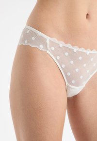 Tanga de encaje blanco con parte delantera transparente con patrones de lunares, bordes festoneados y parte trasera blanca sólida. Textura suave y diseño ligero.