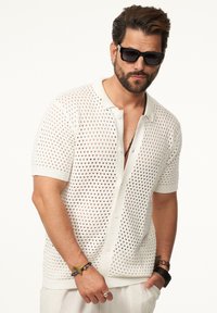 Weißes gestricktes Kurzarmshirt mit offenem Mesh-Design, ausgestattet mit einem Kragen, Knopfverschluss und gerippten Bündchen. Getragen mit Accessoires.