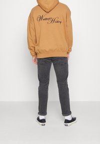 Sweat à capuche marron avec le texte « Written History » dans le dos, associé à un jean noir et des baskets noires. Coupe décontractée et confortable.