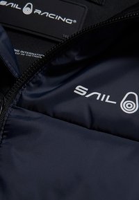 Indersiden af jakken er lavet af marinefarvet stof med en glat tekstur, sort lynlås og hvid "SAIL RACING"-logo. Den indeholder pleje- og mærkningstags.