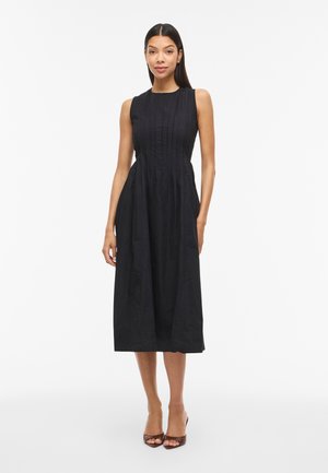 Moteris vilki juodą be rankovių midi ilgumo su klostuotais detalėmis suknią, stovi tiesiai priešais vientisą baltą foną.