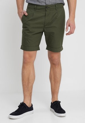 Mand iført opkrogede mørkegrønne shorts, grå skjorte og sorte sneakers med hvide såler, stående på en ensfarvet baggrund.