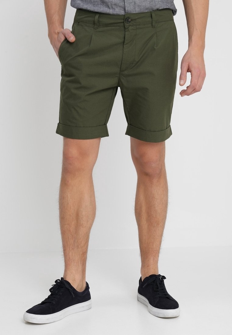 Homme portant un short vert foncé retroussé, une chemise grise et des baskets noires aux semelles blanches, debout sur un fond uni.
