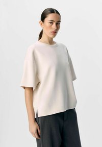Ljus beige översized kortärmad skjorta med rund halsringning och sidofickor, tillverkad av ett mjukt tyg. Kombineras med mörkgrå byxor.