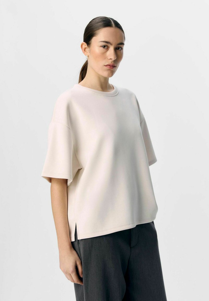 Ljus beige översized kortärmad skjorta med rund halsringning och sidofickor, tillverkad av ett mjukt tyg. Kombineras med mörkgrå byxor.