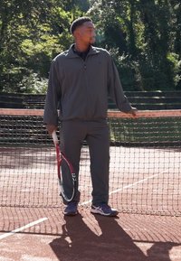 Idrettsutøver i mørkegrå treningsdrakt står ved et tennisnett, holder en rød og svart tennisracket, iført mørke sko, på en leircourt.