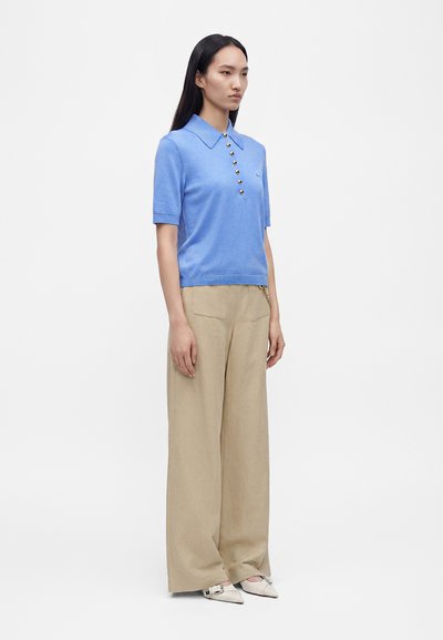 Femme debout portant un polo bleu à manches courtes avec des boutons dorés et un pantalon large beige avec des chaussures blanches pointues.
