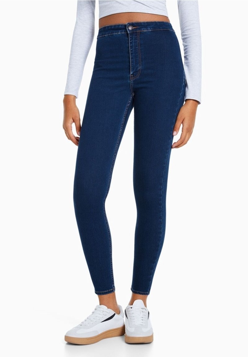 Jeans scuri a vita alta con un design aderente, texture liscia, cuciture visibili e chiusura a bottone, abbinati a sneakers bianche.