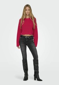 Maglione corto fucsia con texture a coste, abbinato a jeans slim grigio scuro e stivali neri in pelle al ginocchio con tacco quadrato.