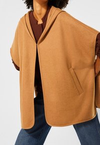 Persona vistiendo una capa con capucha suelta de color camello sobre una camiseta marrón y jeans azules, de pie con las manos en las caderas.