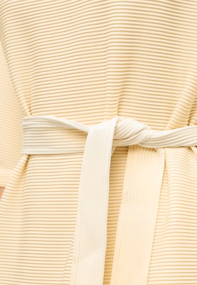 Κοντινό πλάνο μιας ζώνης από ριμπ ύφασμα σε χλωμό μπεζ χρώμα, δεμένη σε κόμπο γύρω από ένδυμα με αντίστοιχη υφή.