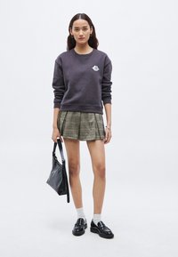 Bluza mov închis cu logo, fustă plisată în carouri, geantă neagră, șosete albe și pantofi negri tip loafer pe un fundal simplu.