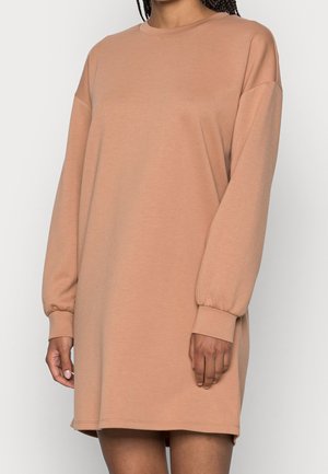 Kobieta ubrana w luźną, długą sukienkę typu sweatshirt w kolorze wielbłądzim z podwiniętymi rękawami, pokazana od szyi do połowy uda na jednolitym tle.
