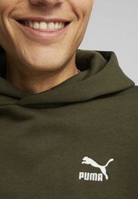 Puma mikina s kapucňou v tmavozelenej farbe s textúrovaným povrchom. Obsahuje biele logo na hrudníku a široký, kapucňový výstrih.