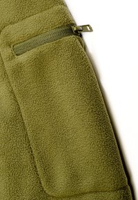 Tissu en polaire vert olive avec une petite poche zippée et un curseur de fermeture éclair visible sur le côté supérieur gauche du vêtement.