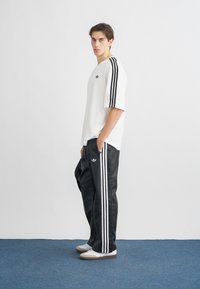White cotton t-shirt with black three-stripe details and logo, paired with black satin jogger pants featuring side stripes and pockets. White shoes.