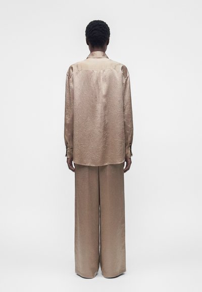 Άτομο με κοντά σγουρά μαλλιά που φοράει μια φαρδιά, λαμπερή μπεζ πουκάμισο και παντελόνια με φαρδιά πόδια, στέκεται μπροστά σε ένα απλό λευκό φόντο.