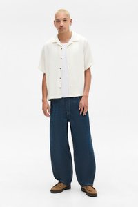 Kortermet kremfarget skjorte med teksturert stoff, over en hvit t-skjorte; kombinert med løse mørkeblå jeans og brune semskede sko.