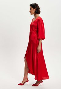 Red satin wrap dress with puff sleeves and asymmetrical hem, featuring a side slit and adjustable tie at the waist. Paired with red heeled sandals.