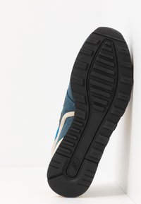 Semelle en caoutchouc noire avec motif de bande de roulement sur une basket bleue, positionnée verticalement contre le coin blanc du mur et du sol.