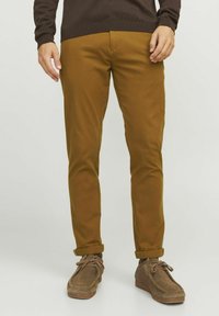 Pantalons en coton marron avec une coupe slim, présentant des poignets roulés. Associés à des chaussures en suède marron. Texture lisse et design décontracté.